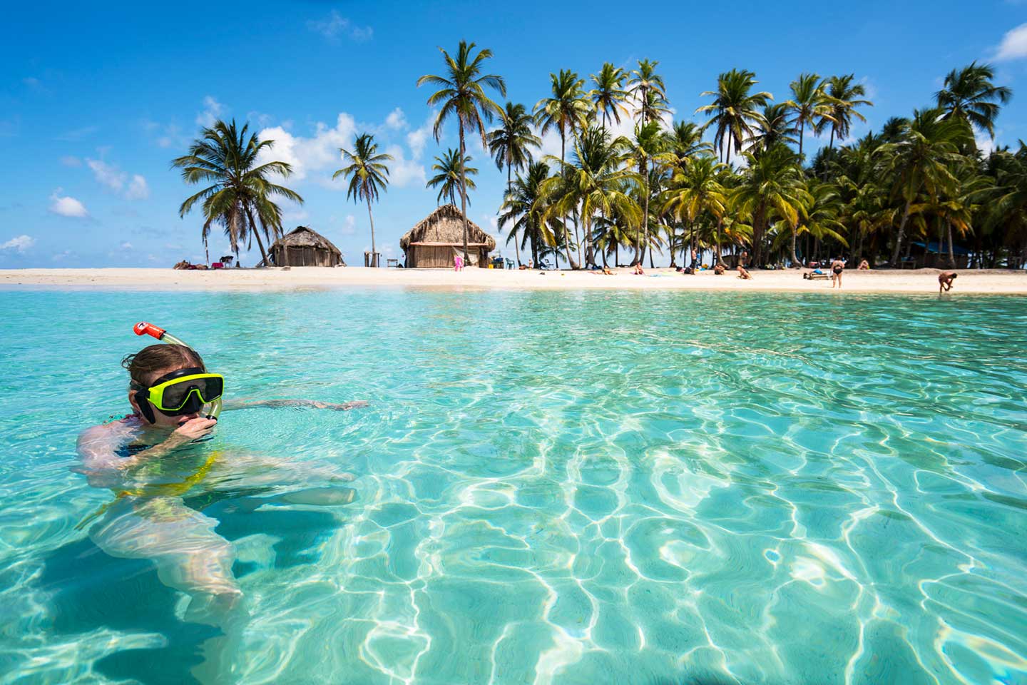 San Blas Inseln - Eine Frau schnorchelt in türkisfarbengrünem Wasser. Im Hintergrund eine kleine Insel mit Palmen und Strohhütten.