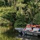 Eine Gruppe von Touristen in orangen Schwimmwesten sitzt in einem langen Boot, das auf einem ruhigen Fluss in Costa Rica fährt, umgeben von dichtem, grünem Regenwald, im Rahmen einer zweiwöchigen Gruppenreise.